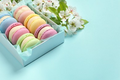 Photo of Many delicious colorful macarons in box and flowers on light blue background, space for text