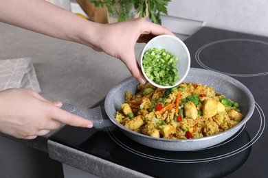 Woman adding cut green onion to rice with meat and vegetables in frying pan, closeup
