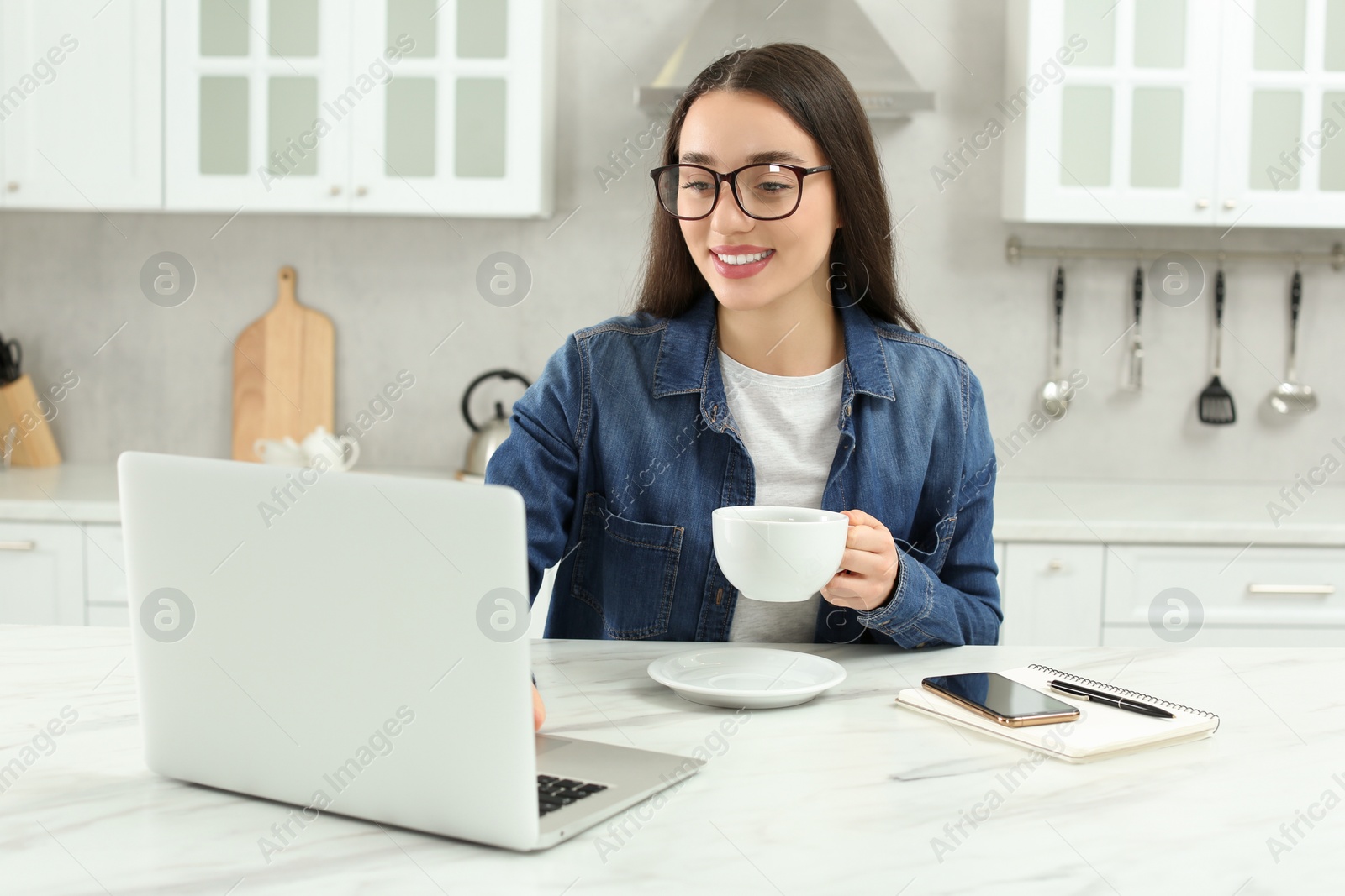 Photo of Home workplace. Happy woman with cup of hot drink looking at laptop at marble desk in kitchen