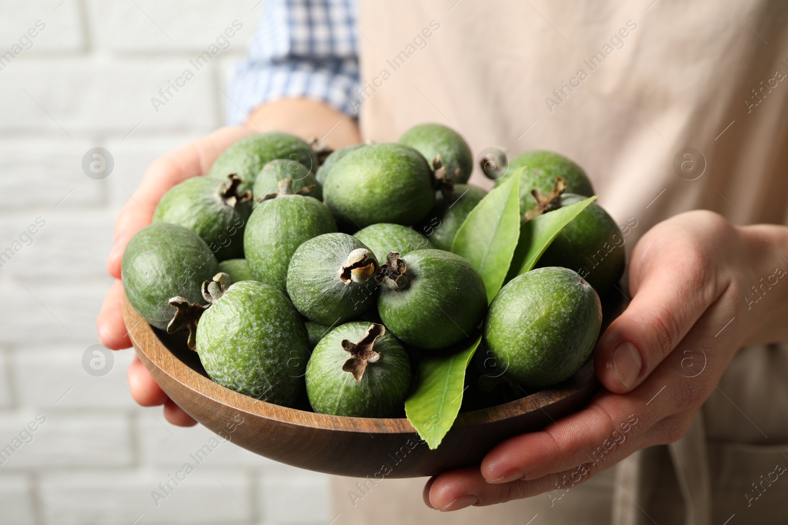 Photo of Woman holding fresh green feijoa fruits in bowl against white brick wall, closeup
