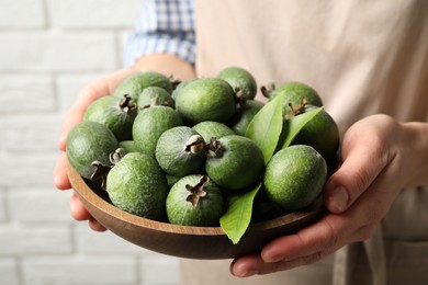 Woman holding fresh green feijoa fruits in bowl against white brick wall, closeup