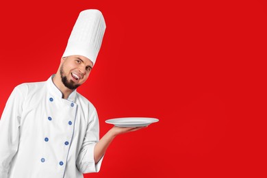 Photo of Happy professional confectioner in uniform holding empty plate on red background. Space for text