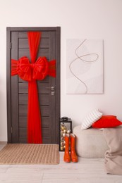 Wooden door with beautiful bow near container with baubles, ottoman and red rubber boots in room. Christmas decoration