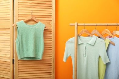 Photo of Sweater vest hanging on wooden folding screen and rack with stylish women's clothes near orange wall