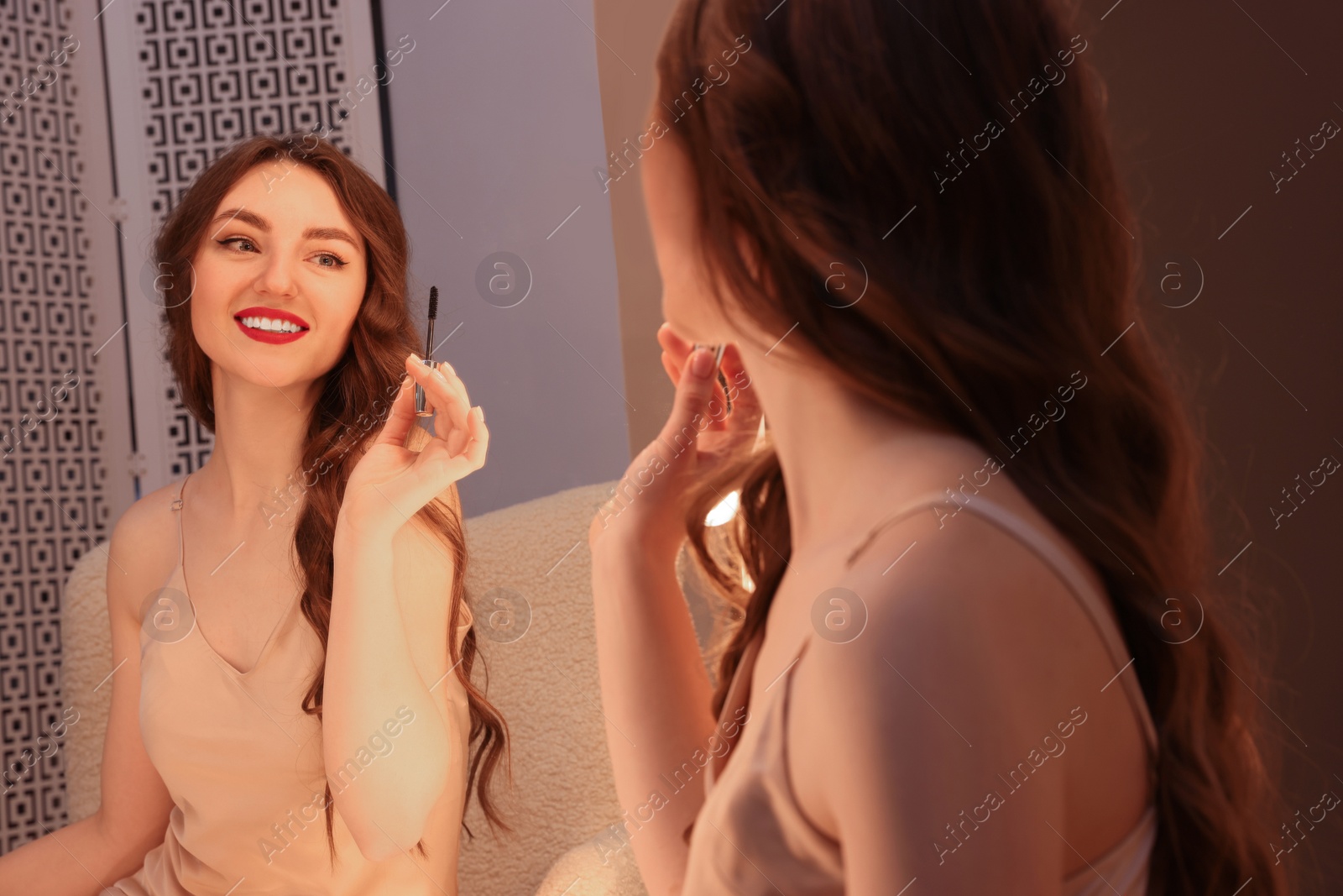 Photo of Beautiful young woman in elegant dress with mascara brush near mirror indoors
