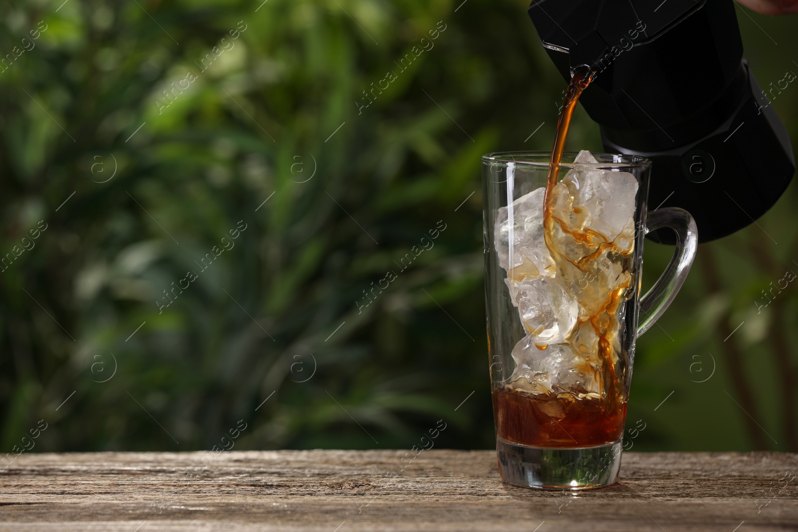 Photo of Pouring coffee over ice in glass cup at wooden table outdoors. Space for text