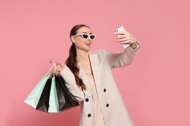 Photo of Stylish young woman with shopping bags taking selfie on pink background