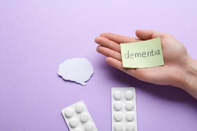 Photo of Woman holding paper note with word Dementia near pills and brain cutout on violet background, top view