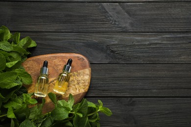 Photo of Bottles of mint essential oil and green leaves on black wooden table, flat lay. Space for text