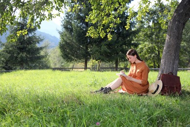 Photo of Beautiful young woman drawing with pencil in notepad near tree on green grass