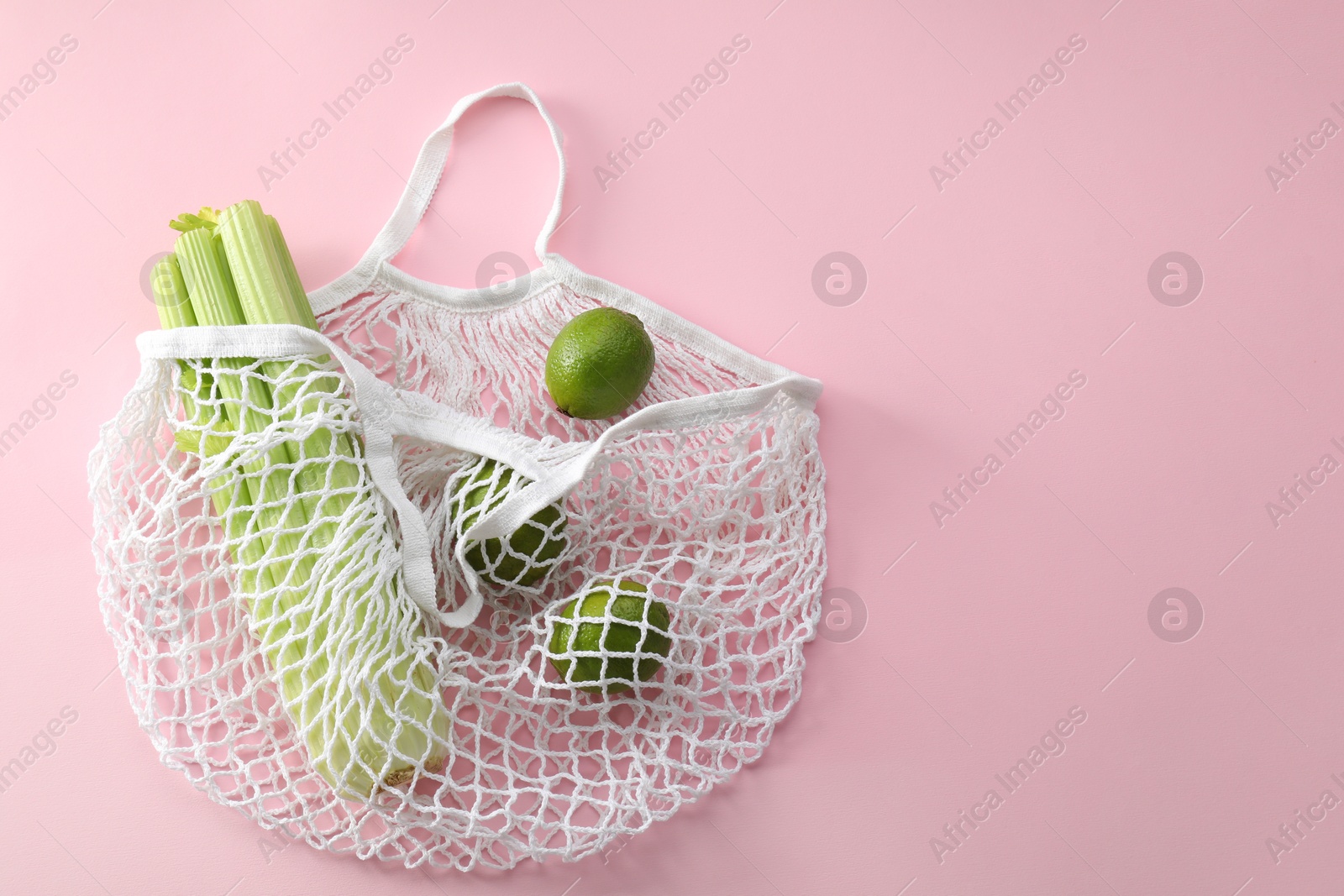 Photo of String bag with celery and limes on pink background, top view. Space for text