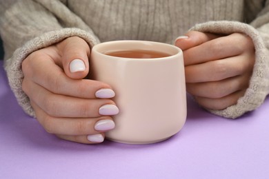 Photo of Woman with white polish on nails holding cup of hot drink on violet background, closeup