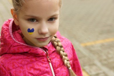 Little girl with drawing of Ukrainian flag on face in heart shape outdoors, space for text
