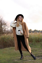 Photo of Beautiful young woman with cup of coffee wearing stylish autumn clothes outdoors