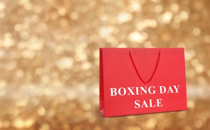 Red shopping bag with text Boxing Day Sale on blurred gold background, closeup. Space for text
