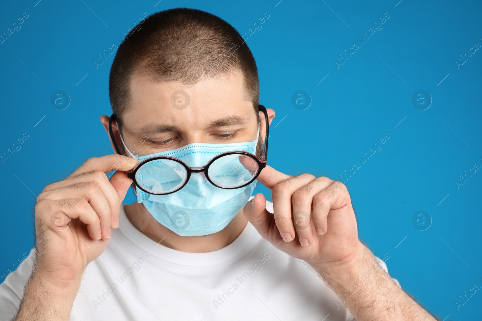 Photo of Man wiping foggy glasses caused by wearing disposable mask on blue background. Protective measure during coronavirus pandemic