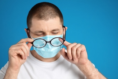 Photo of Man wiping foggy glasses caused by wearing disposable mask on blue background. Protective measure during coronavirus pandemic