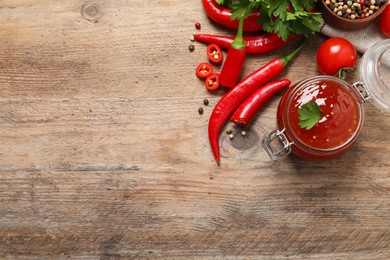 Photo of Spicy chili sauce in glass jar and ingredients on wooden table, flat lay