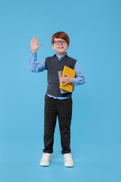 Smiling schoolboy with books waving hello on light blue background