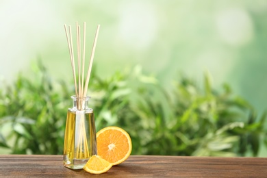 Photo of Reed air freshener and orange on wooden table against blurred green background. Space for text