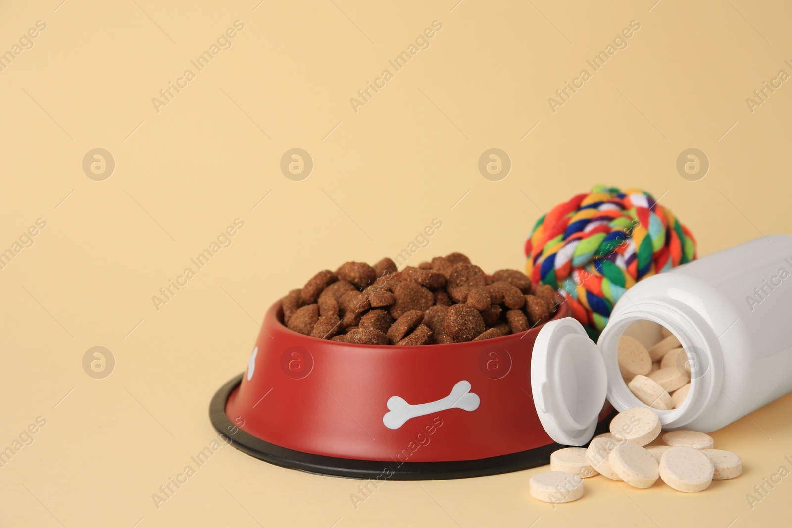 Photo of Bottle with vitamins, dry pet food in bowl and toy on beige background, space for text