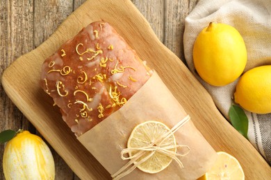 Photo of Wrapped tasty lemon cake with glaze and citrus fruits on wooden table, flat lay