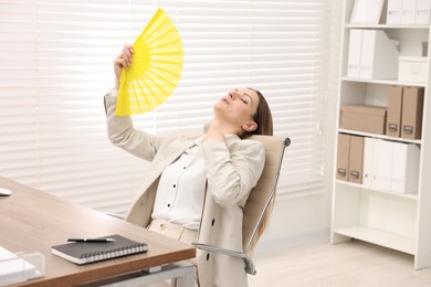 Photo of Businesswoman waving yellow hand fan to cool herself at table in office