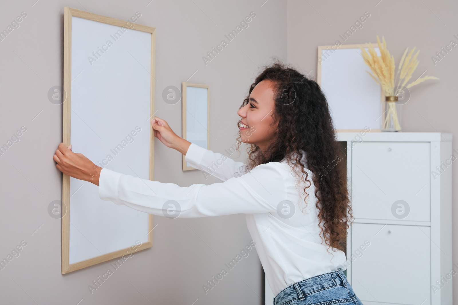Photo of Happy African American woman hanging empty frame on pale rose wall in room. Mockup for design