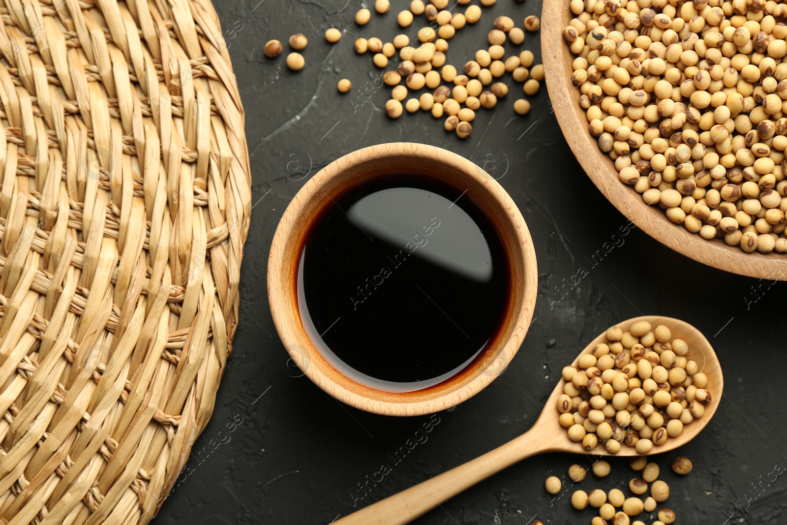 Photo of Soy sauce in bowl and soybeans on black textured table, flat lay