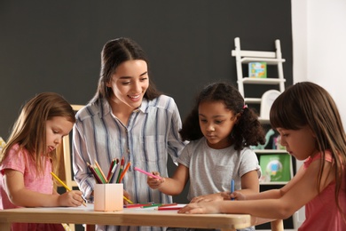 Cute little children with nursery teacher drawing at table in kindergarten. Indoor activity