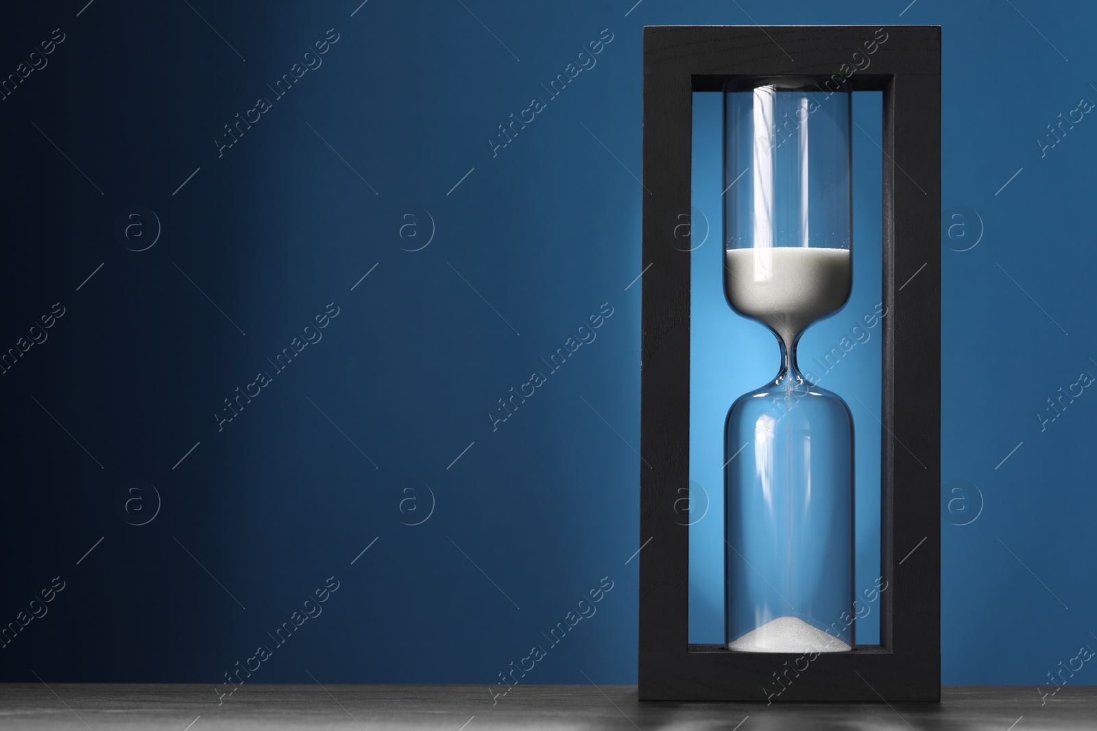 Photo of Hourglass with flowing sand on table against blue background, space for text