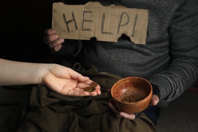 Photo of Woman giving coins to poor homeless man with bowl of donations and help sign on dark background, closeup