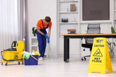 Photo of Cleaning service worker washing floor with mop, selective focus. Bucket with supplies and wet floor sign in office