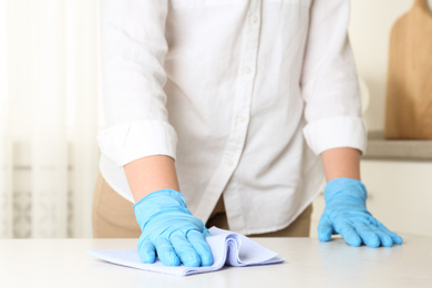 Photo of Woman in gloves wiping white table with rag indoors, closeup