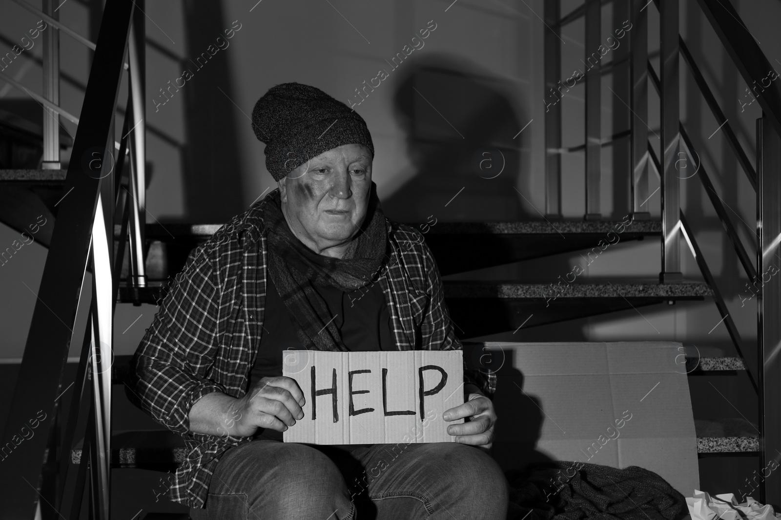 Photo of Poor senior man with cardboard sign HELP on stairs indoors. Black and white effect