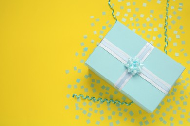 Light blue gift box, streamers and confetti on yellow background, top view. Space for text