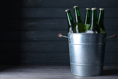 Metal bucket with bottles of beer and ice cubes on wooden table against dark background, space for text