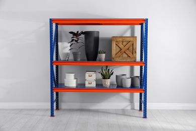 Photo of Metal shelving unit with different household stuff near light wall indoors
