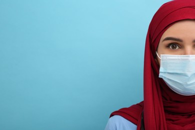 Photo of Muslim woman wearing hijab, medical uniform and protective mask on light blue background, closeup. Space for text
