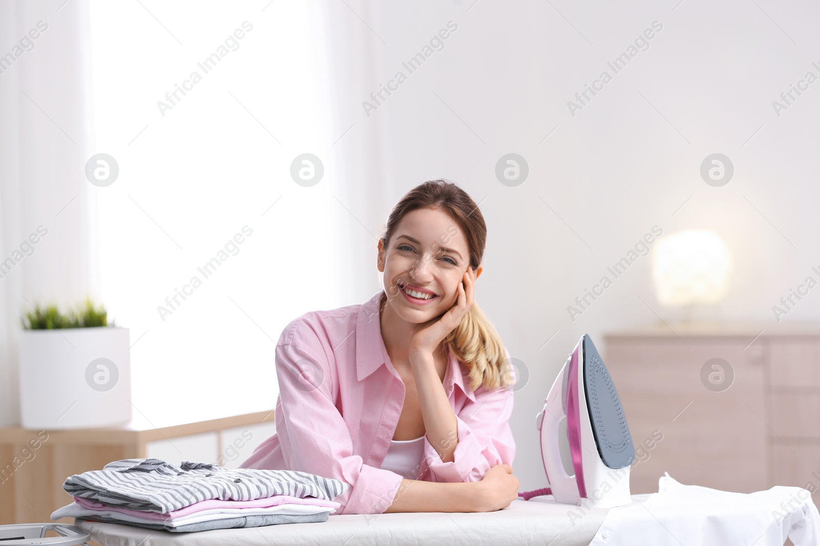 Photo of Young pretty woman with iron and clean laundry at board indoors