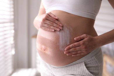 Photo of Young pregnant woman applying cosmetic product on belly indoors, closeup