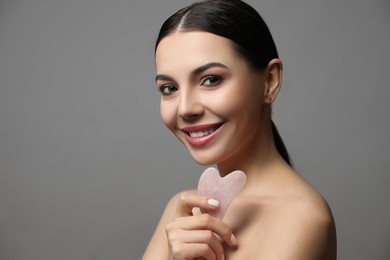 Beautiful young woman doing facial massage with gua sha tool on grey background. Space for text