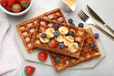 Photo of Delicious Belgian waffles with banana, berries and caramel sauce on light marble table, flat lay