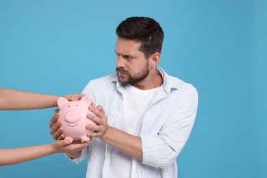 Photo of Woman taking piggy bank from man on light blue background. Be careful - fraud
