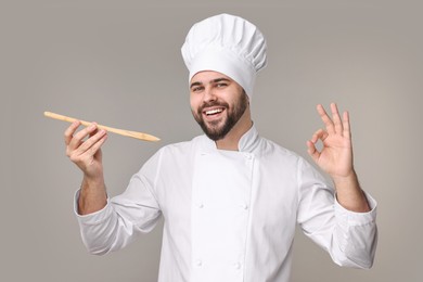 Photo of Happy young chef in uniform holding wooden spoon and showing ok gesture on grey background