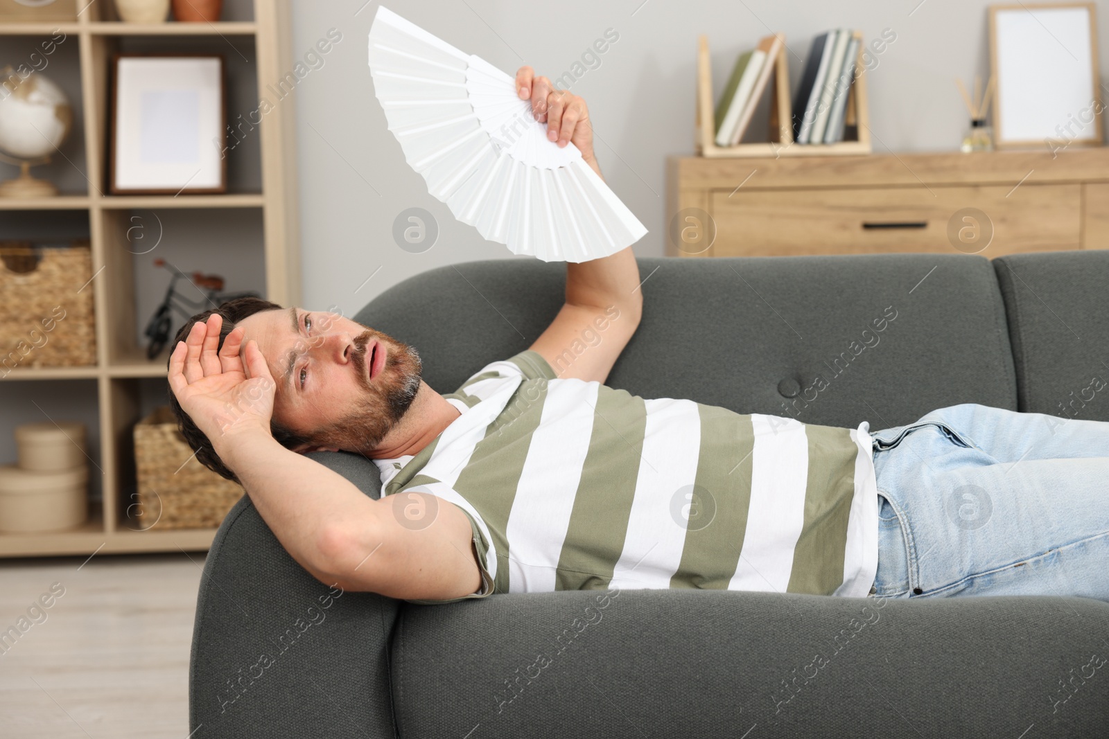 Photo of Bearded man waving white hand fan to cool himself on sofa at home