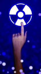 Woman touching icon of glowing radiation warning symbol on blue background with blurred lights, closeup