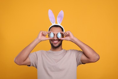 Photo of Happy African American man in bunny ears headband covering eyes with Easter eggs on orange background