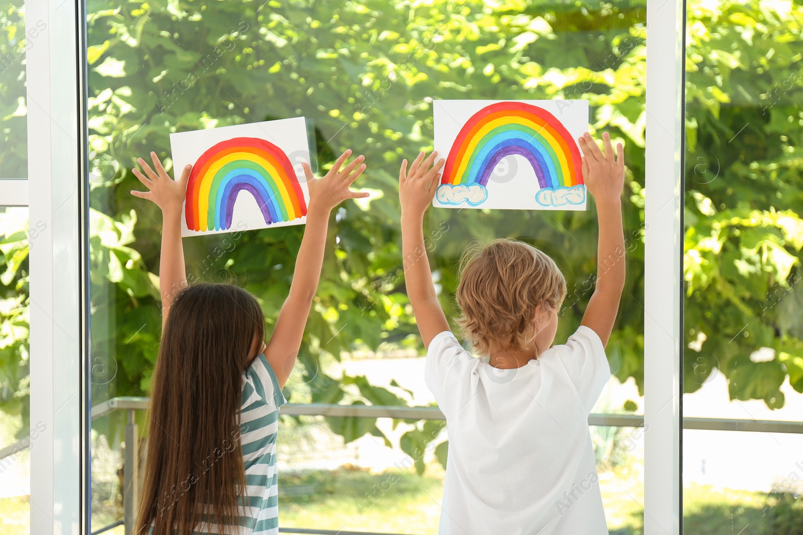 Photo of Little children holding rainbow paintings near window indoors, back view. Stay at home concept