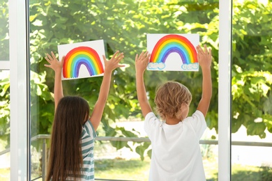 Photo of Little children holding rainbow paintings near window indoors, back view. Stay at home concept